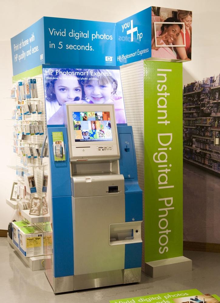 This touchscreen kiosk for HP's Photosmart Express is wrapped in signage to attract users in locations such as supermarkets and pharmacies. Note how the signs peek above the height of the shelves, and there is even a mat on the aisle floor. Courtesy Hewlett-Packard.