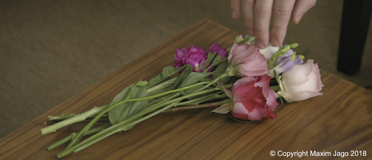 A screenshot depicts image of a bunch of flowers on a table with the finger tips of a person on the flowers.