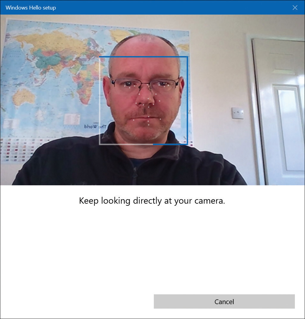 This screenshot shows the Windows Hello setup window. In the center of the window is a picture of a face with several dots or markers overlaid on the image. Below the image, the text Keep Looking Directly At Your Camera appears. Below this text is a Cancel button.