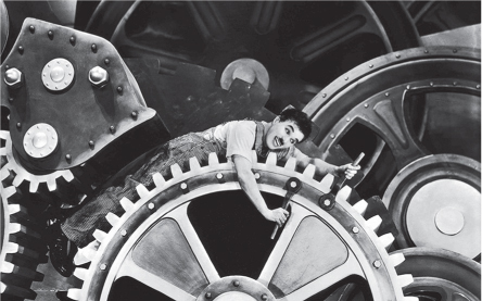 A still from the movie Little Tramp shows giant gears connected to other mechanical parts, and Charlie Chaplin lying on one of the gears and wrenching two screws, each with one hand.