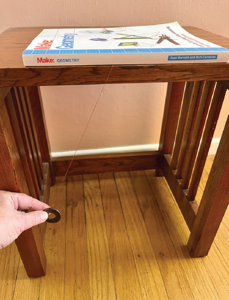 The piece of string with is now hanging from the edge of a table, with the second washer held down with a book to anchor it. A hand is holding the first washer up to one side with the string taut, ready to release it to swing freely.